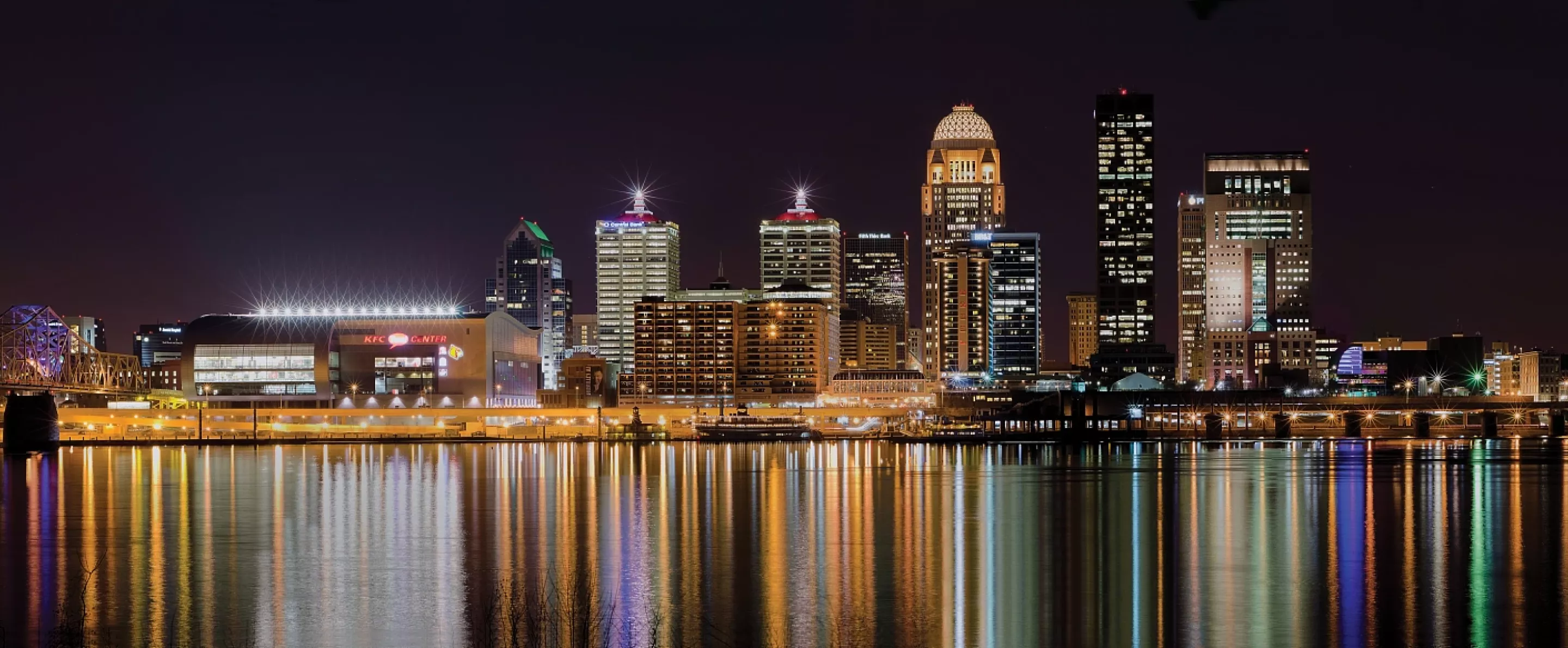 Darker nighttime skyline photo of the Louisville waterfront with light reflections in the Ohio River