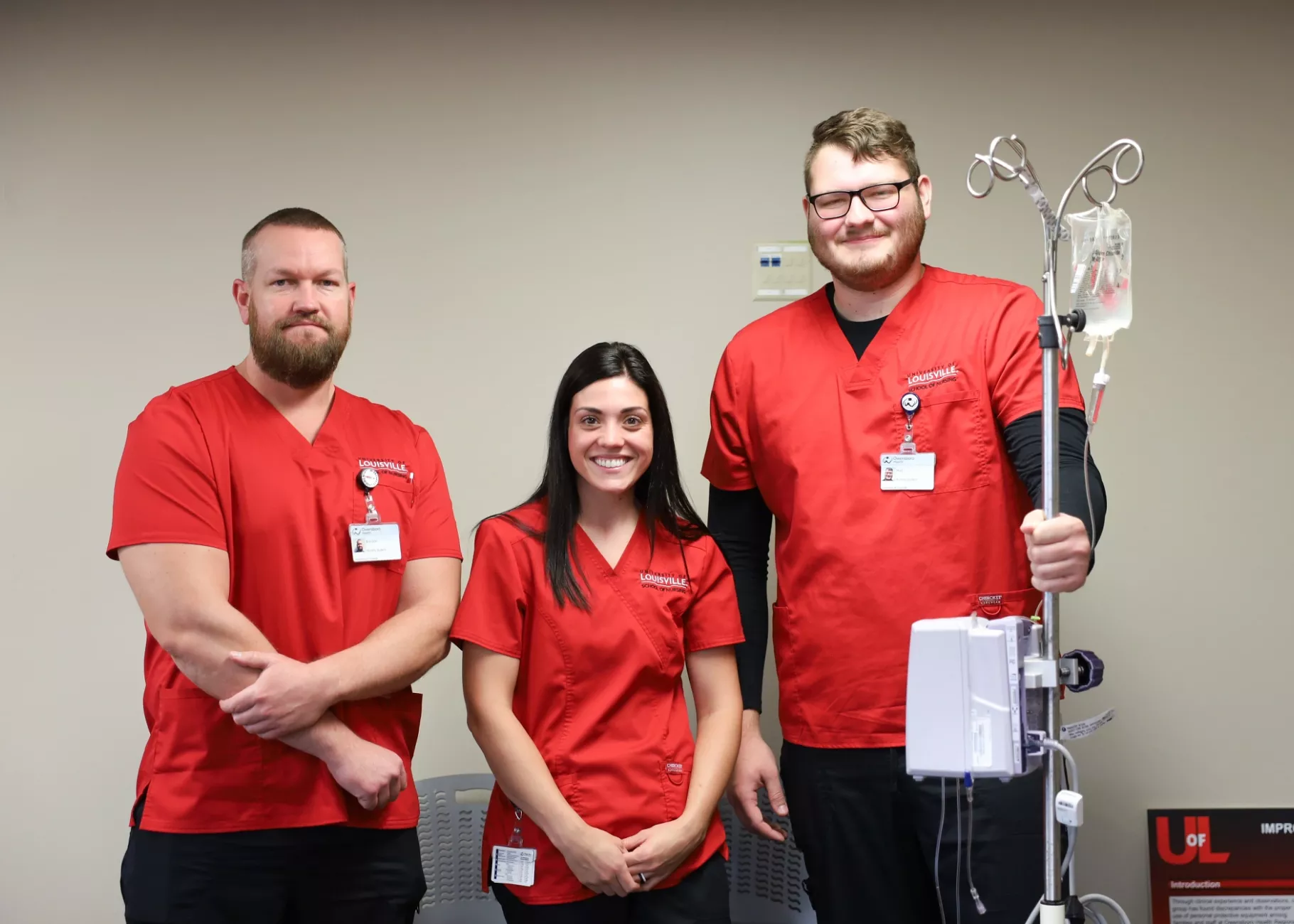 University of Louisville School of Nursing Owensboro Extension. Three students in red scrubs stand next to an IV pump in the classroom.