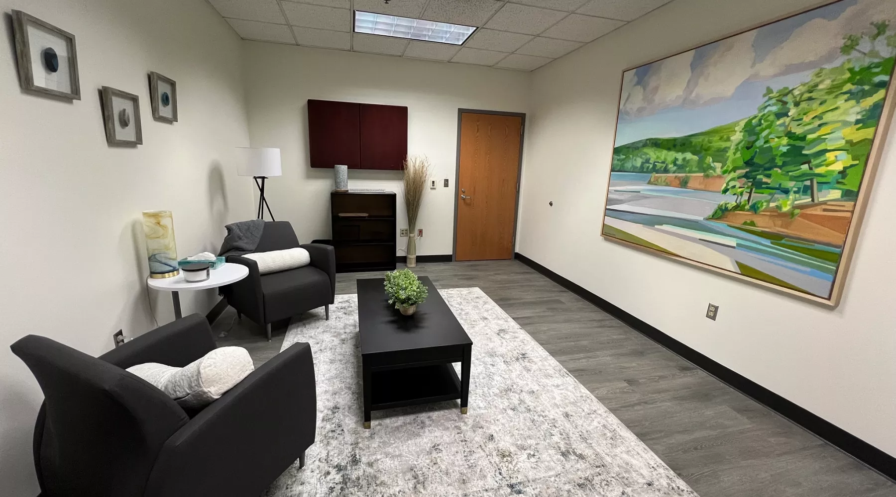 Wellness room in the University of Louisville School of Nursing 4th floor on HSC campus. The room is spacious and has minimal furniture. A large painting of a landscape covers one wall. Two chairs and a coffee table sit in the middle of the room.