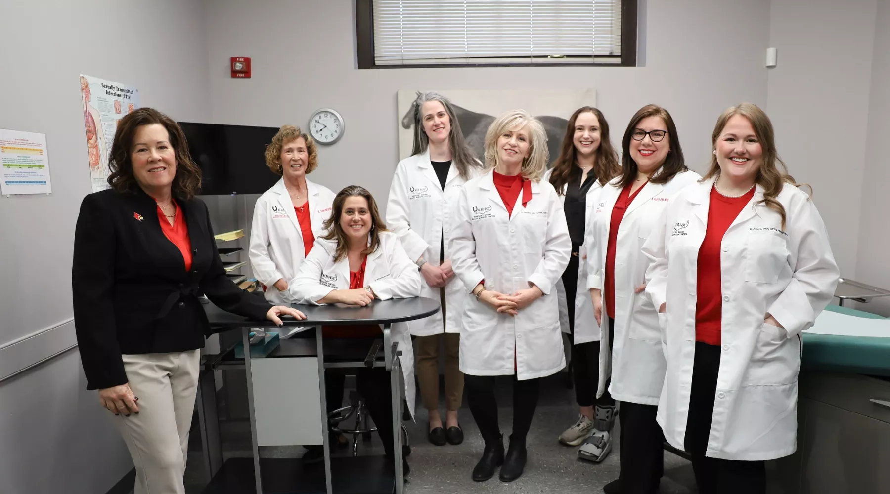 Kentucky Racing Health Services Center (KRHSC) Staff and University of Louisville School of Nursing faculty in an exam room located in the Center. A photo of a horse is behind the members. They are wearing their white lab coats.