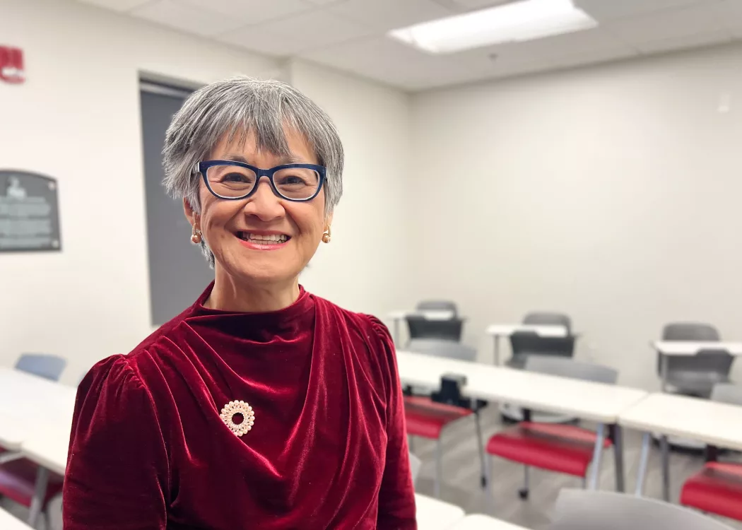 UofL School of Nursing Professor Dr. Ratchneewan Ross smiling at the camera in a classrrom. Student rows fo desks and chairs are behind her.