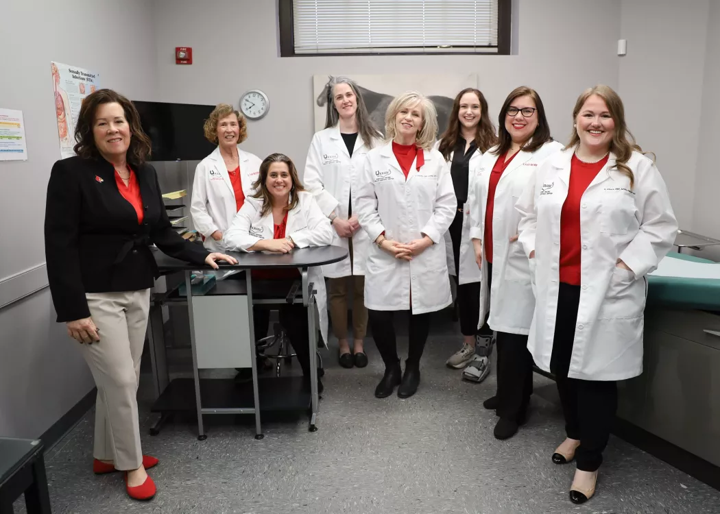 Kentucky Racing Health Services Center (KRHSC) Staff and University of Louisville School of Nursing faculty in an exam room located in the Center. A photo of a horse is behind the members. They are wearing their white lab coats.
