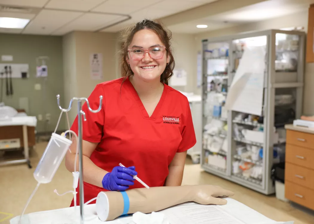 University of Louisville School of Nursing Owensboro Extension. ABSN students practice IV medication administration in the high-fidelity simulation lab. The student smiles at the camera in front of the simulation equipment.