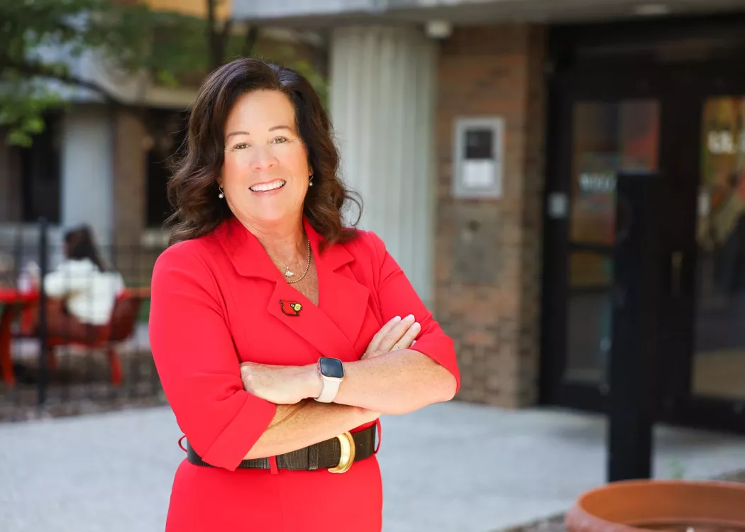 University of Louisville School of Nursing Dean, Dr. Whitney Nash. Nash stands in front of the School of Nursing on HSC campus in a red dress with a cardinal head pin.