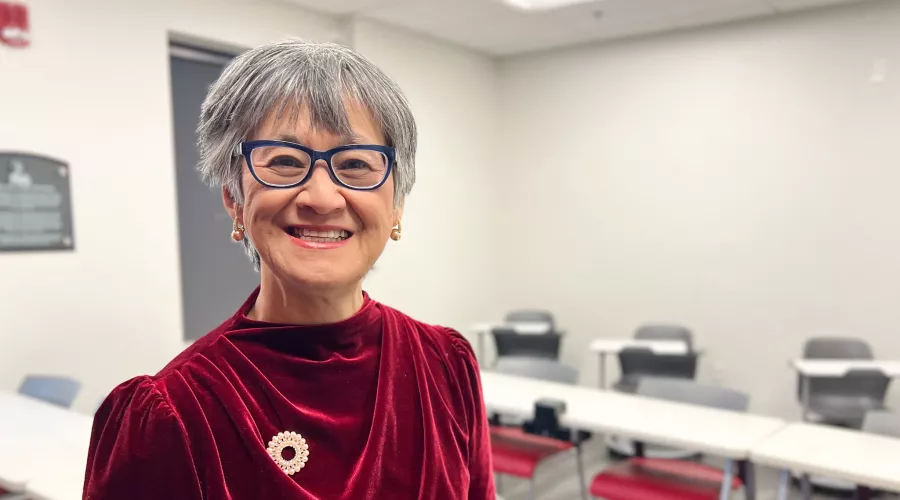 UofL School of Nursing Professor Dr. Ratchneewan Ross smiling at the camera in a classrrom. Student rows fo desks and chairs are behind her.